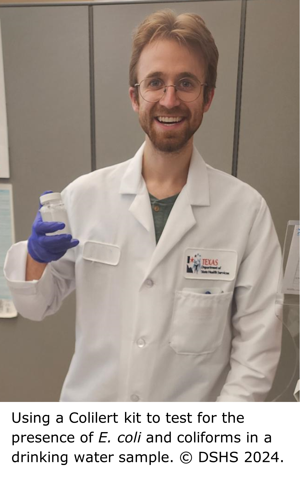 A smiling person in a white coat and wearing gloves holds a small water sample container in their right hand