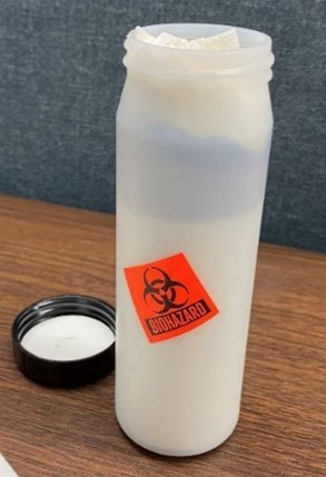 "A photo of a plastic cylindrical secondary container with a black lid. The container and lid are placed on a table. An orange biohazard sticker is stuck to the side of the container. "
